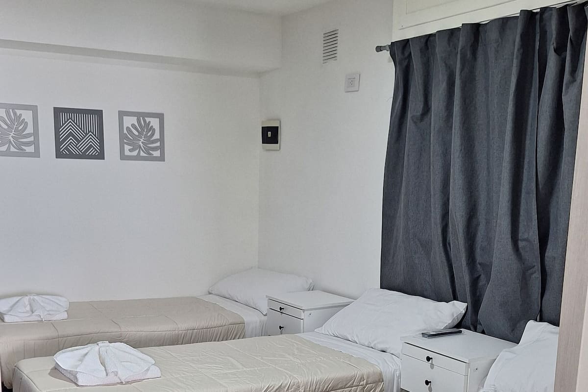 Ein Hotelzimmer mit drei Betten, jedes mit weißer Daunendecke und einem Handtuch darauf. Ein Fenster mit dunklen Vorhängen, eine weiße Wand mit drei gerahmten Bildern und eine Deckenleuchte.