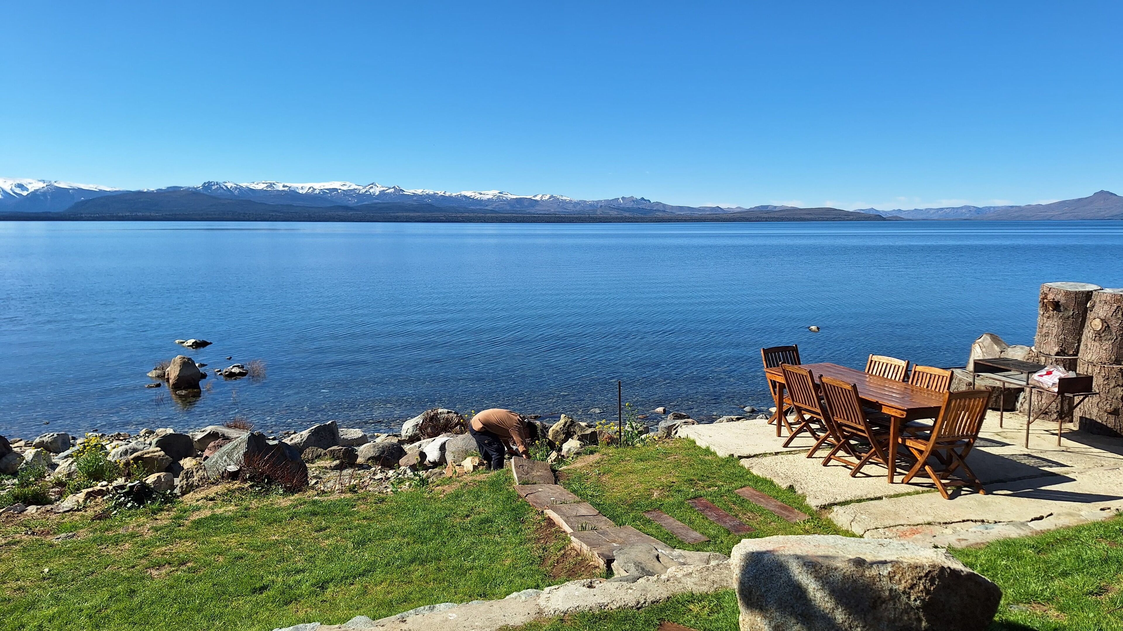 A lakeside seating area with a table and chairs, surrounded by rocks and grass, overlooking a calm lake with mountains in the distance.