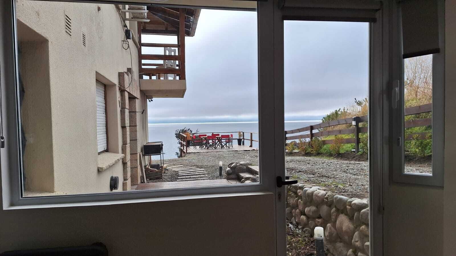 A view from a room with a window overlooking a beach area with a wooden deck and outdoor furniture.