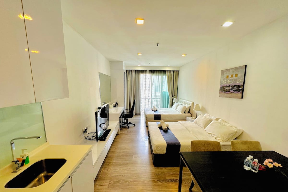 Une chambre d’hôtel avec une kitchenette, deux lits, un bureau et une grande fenêtre avec des rideaux.
