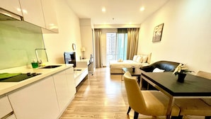 Een moderne hotelkamer met een kitchenette, een eethoek met tafel en stoelen, een bank en een groot raam met gordijnen.
