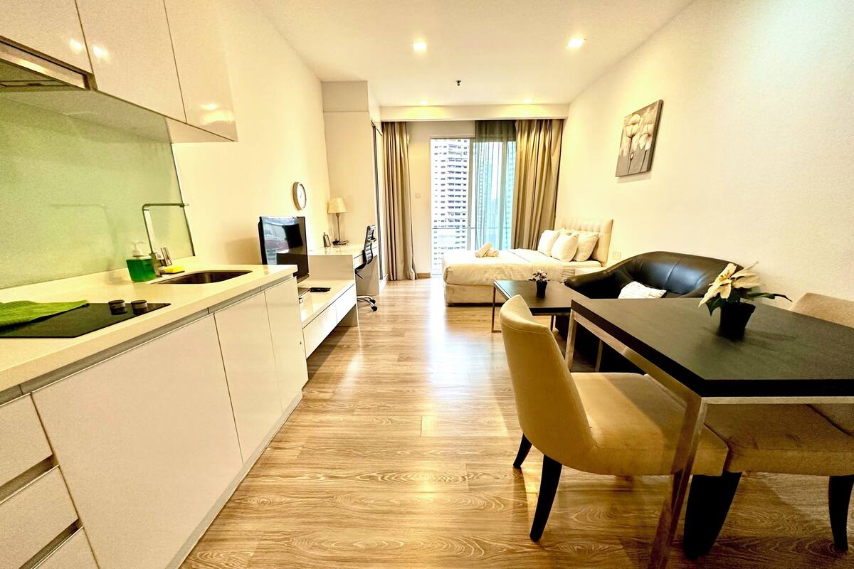 Une chambre d’hôtel moderne dotée d’une kitchenette, d’un coin repas avec une table et des chaises, d’un canapé et d’une grande fenêtre avec des rideaux.