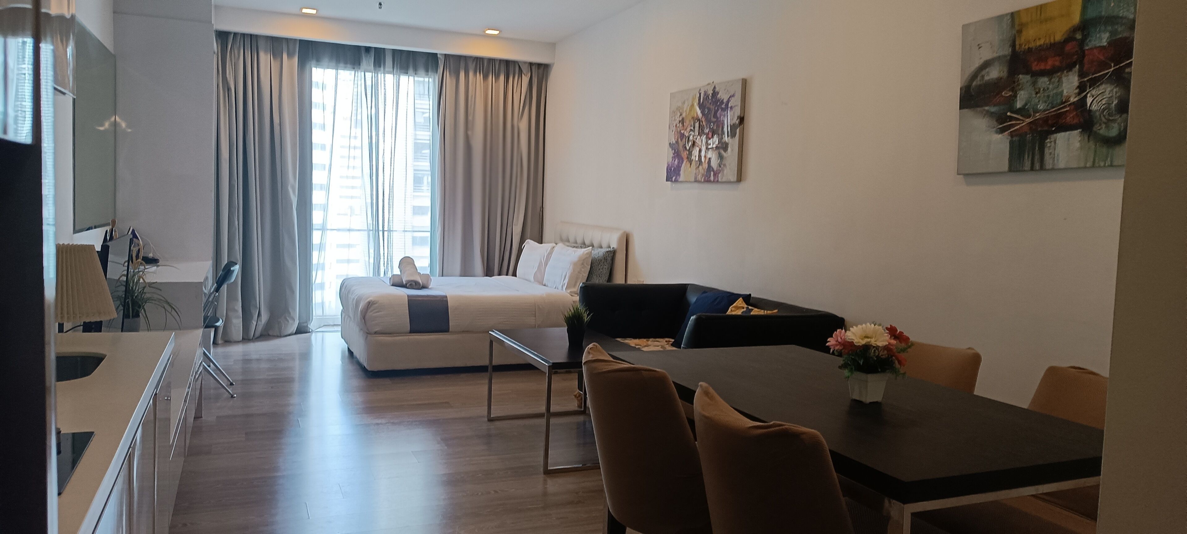 A modern hotel room with a bed, a dining table with chairs, a small coffee table, and a large window with curtains.