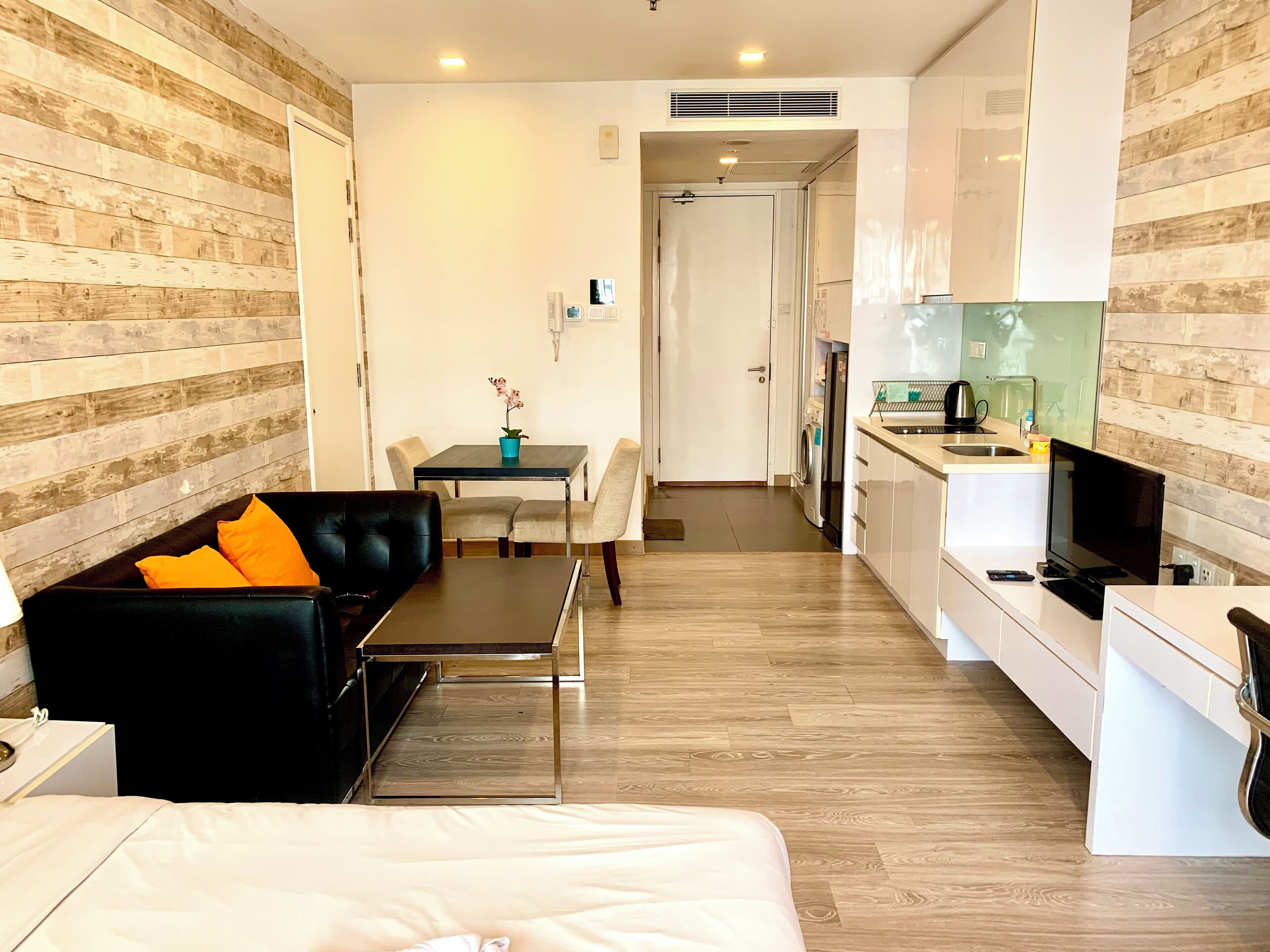 A modern hotel room with a bed, a black leather sofa with orange cushions, a small dining table with chairs, a kitchenette, and a wooden wall paneling.