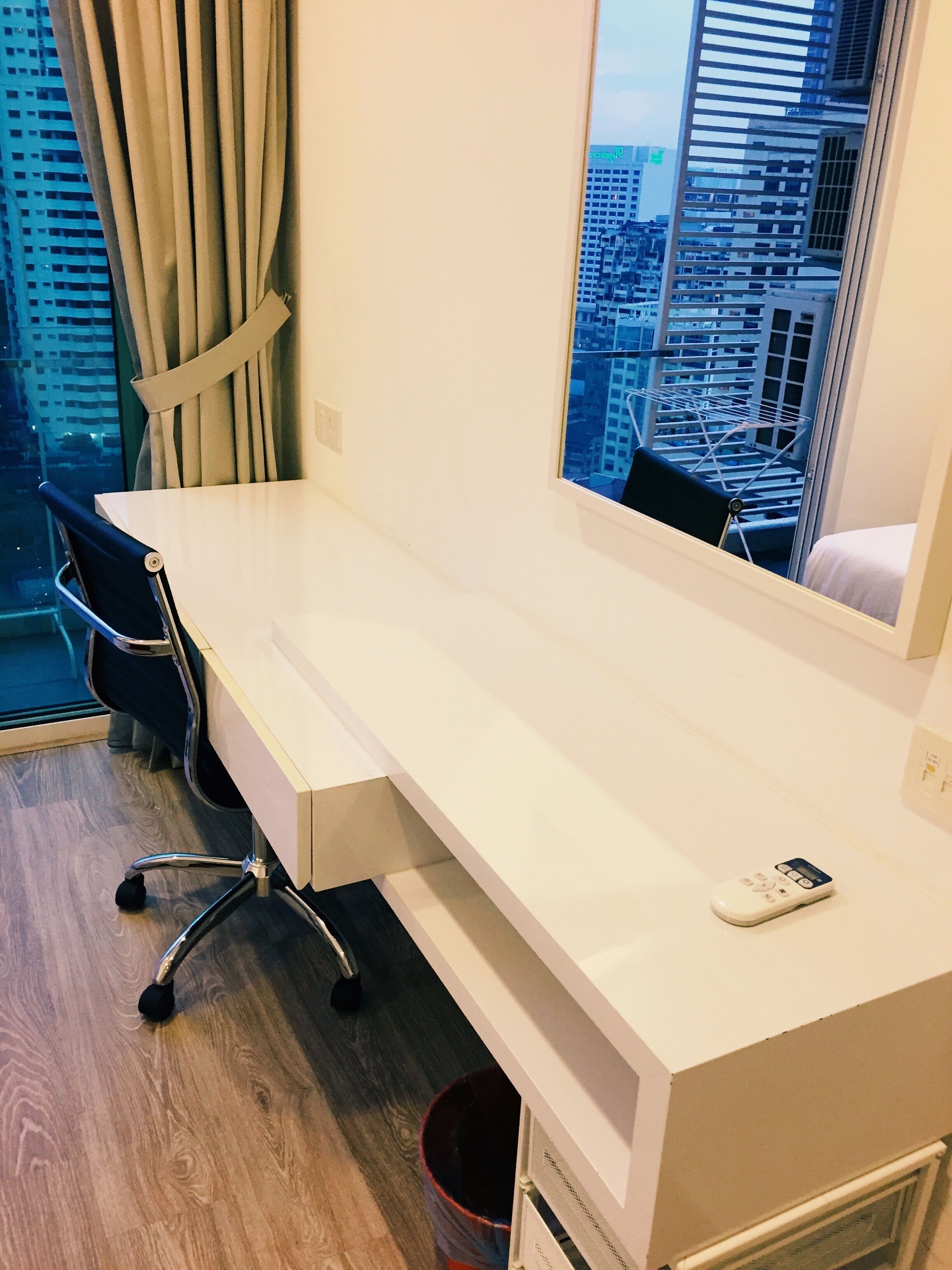 A hotel room with a long white desk, a chair, a small trash bin, and a window with a view of a cityscape.