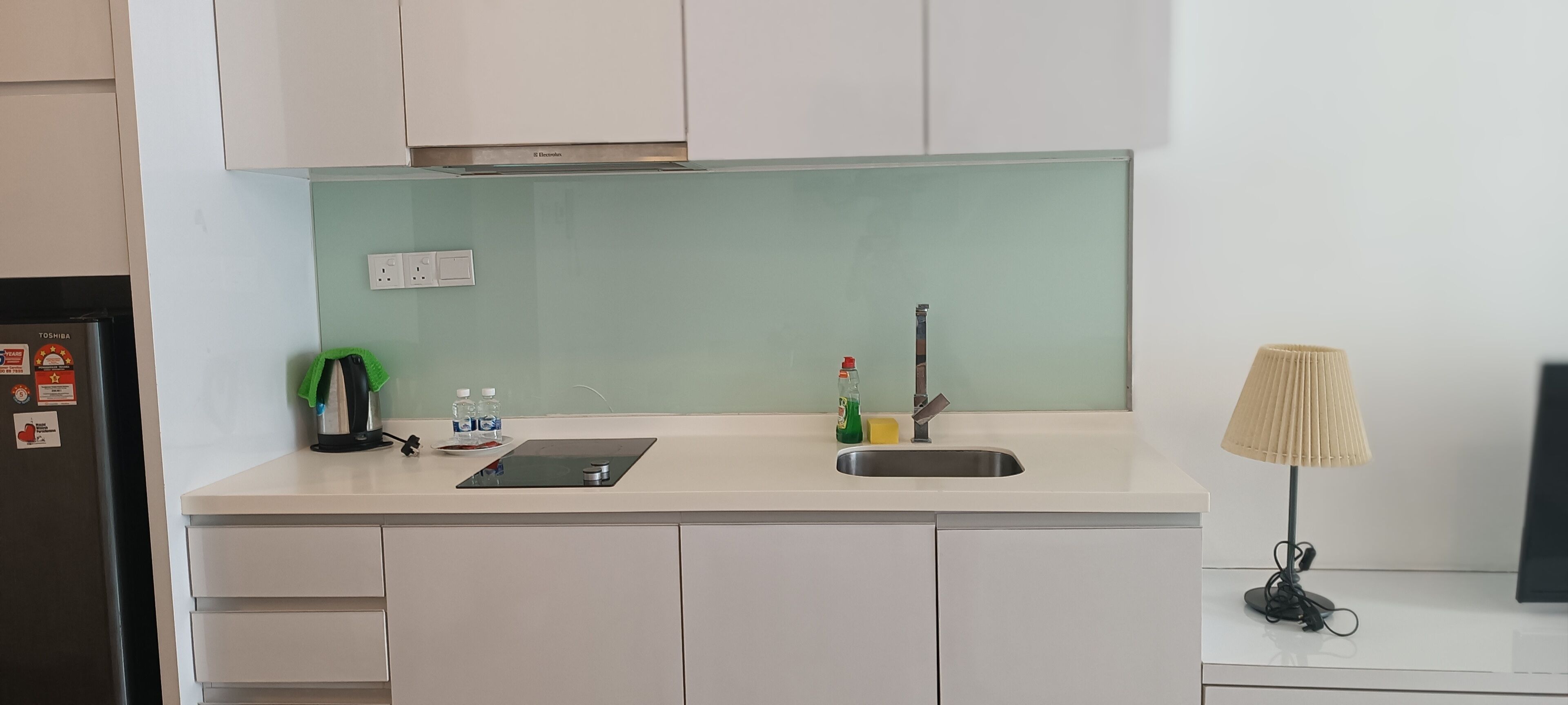 A kitchen with white cabinets, a stainless steel sink, a green countertop, a black refrigerator, a small table with a lamp, and a few kitchen items.