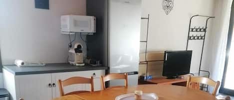 A dining area with a round wooden table, six chairs, a television, and a kitchen area with a refrigerator and microwave.