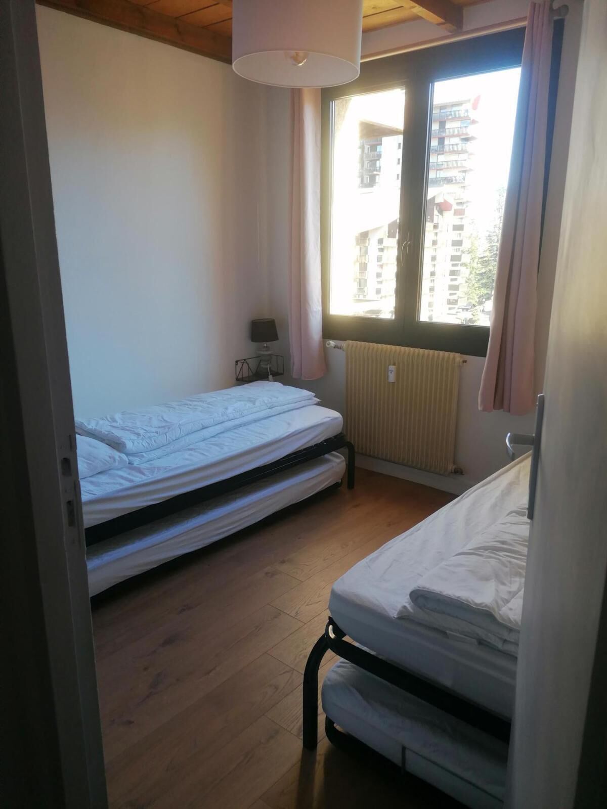 Un petit logement composé d’une seule pièce, comprenant deux lits, une fenêtre avec des rideaux, un radiateur et une lampe sur une table de chevet.