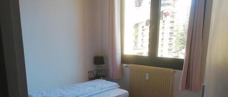 Un petit logement composé d’une seule pièce, comprenant deux lits, une fenêtre avec des rideaux, un radiateur et une lampe sur une table de chevet.