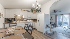 A modern kitchen with white cabinets, a built-in microwave, and a coffee maker. There's a dining area with a table and chairs, and a glassware cart.