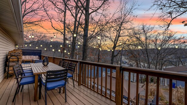 A wooden deck with a table and chairs, string lights, and a view of trees and a sunset.
