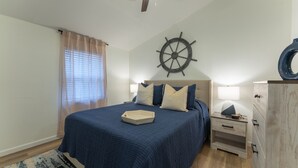 A bedroom with a bed, a nightstand, a dresser, a ship's wheel decoration, and a window with curtains.