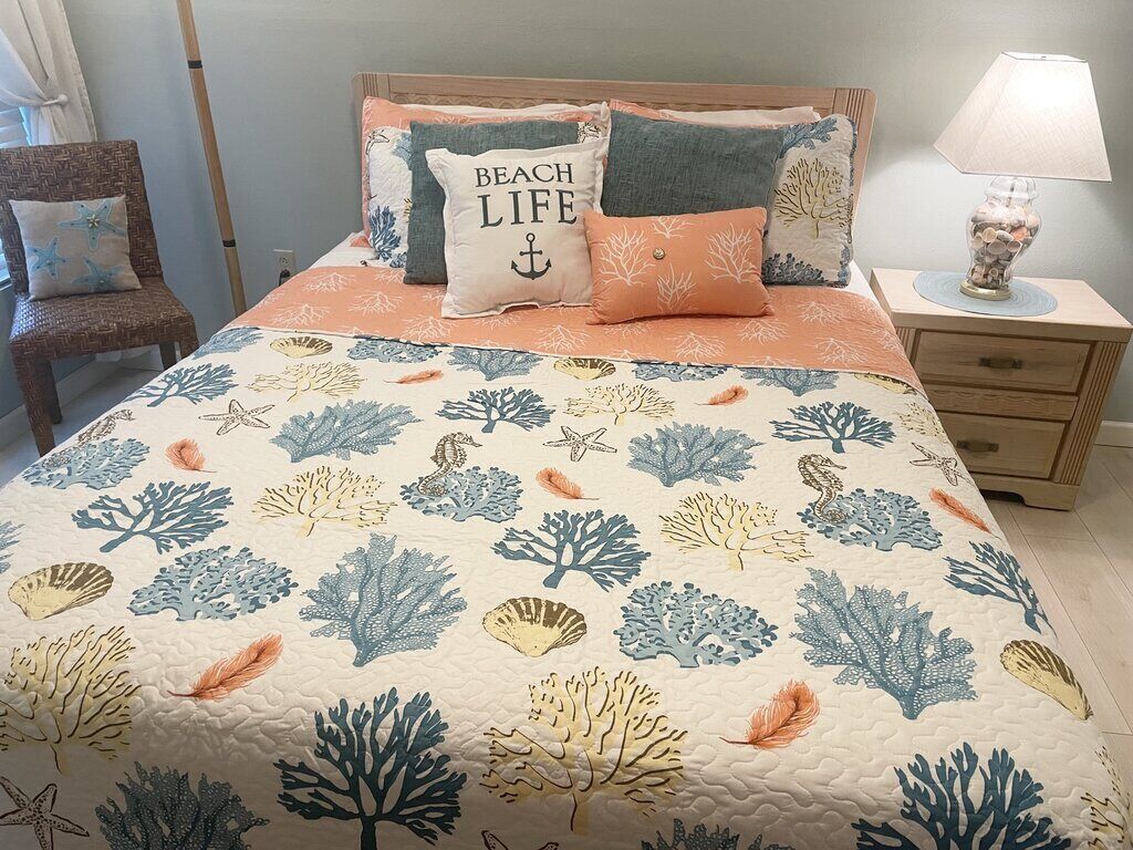 Un dormitorio con una cama adornada con un edredón de temática playera, una mesita de noche de madera con una lámpara y una silla de mimbre.