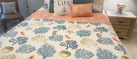 Un dormitorio con una cama adornada con un edredón de temática playera, una mesita de noche de madera con una lámpara y una silla de mimbre.