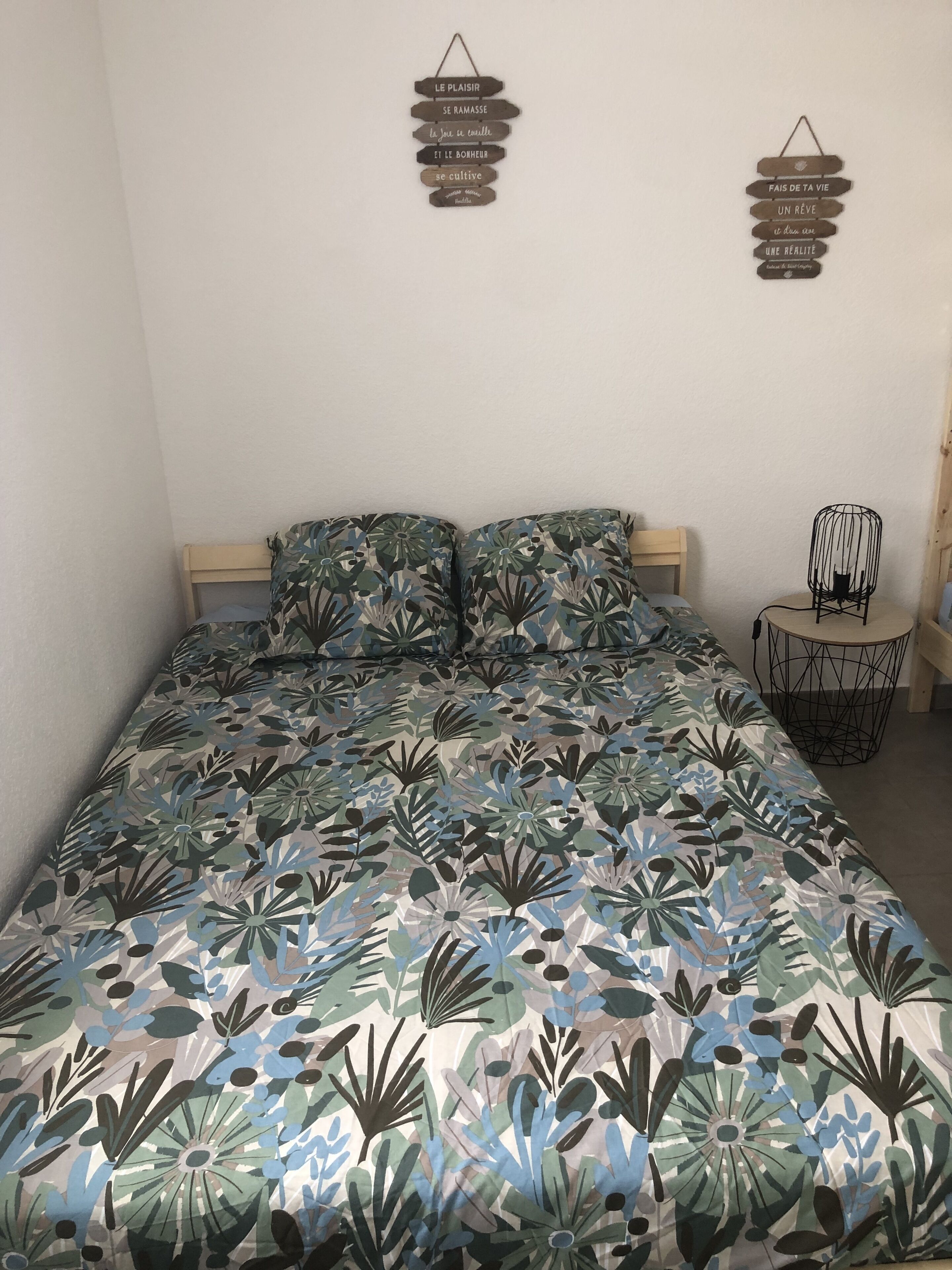 Quarto com colcha estampada de flores, cabeceira de madeira e dois letreiros fixados na parede com texto em francês.