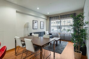 Un salon moderne avec un canapé foncé, une table à manger en bois et des chaises. On y trouve une grande fenêtre avec des rideaux, un lampadaire et une plante en pot.
