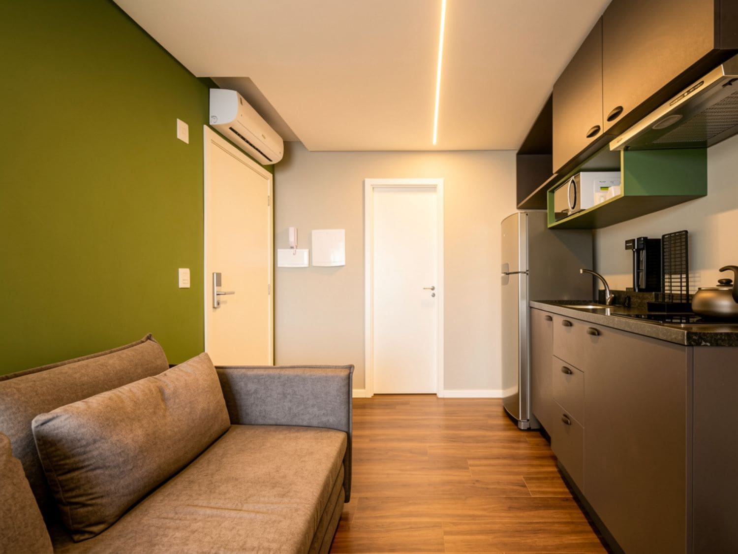 A compact kitchen with a built-in oven, microwave, and a stainless steel refrigerator. There is a wooden floor and a green wall.