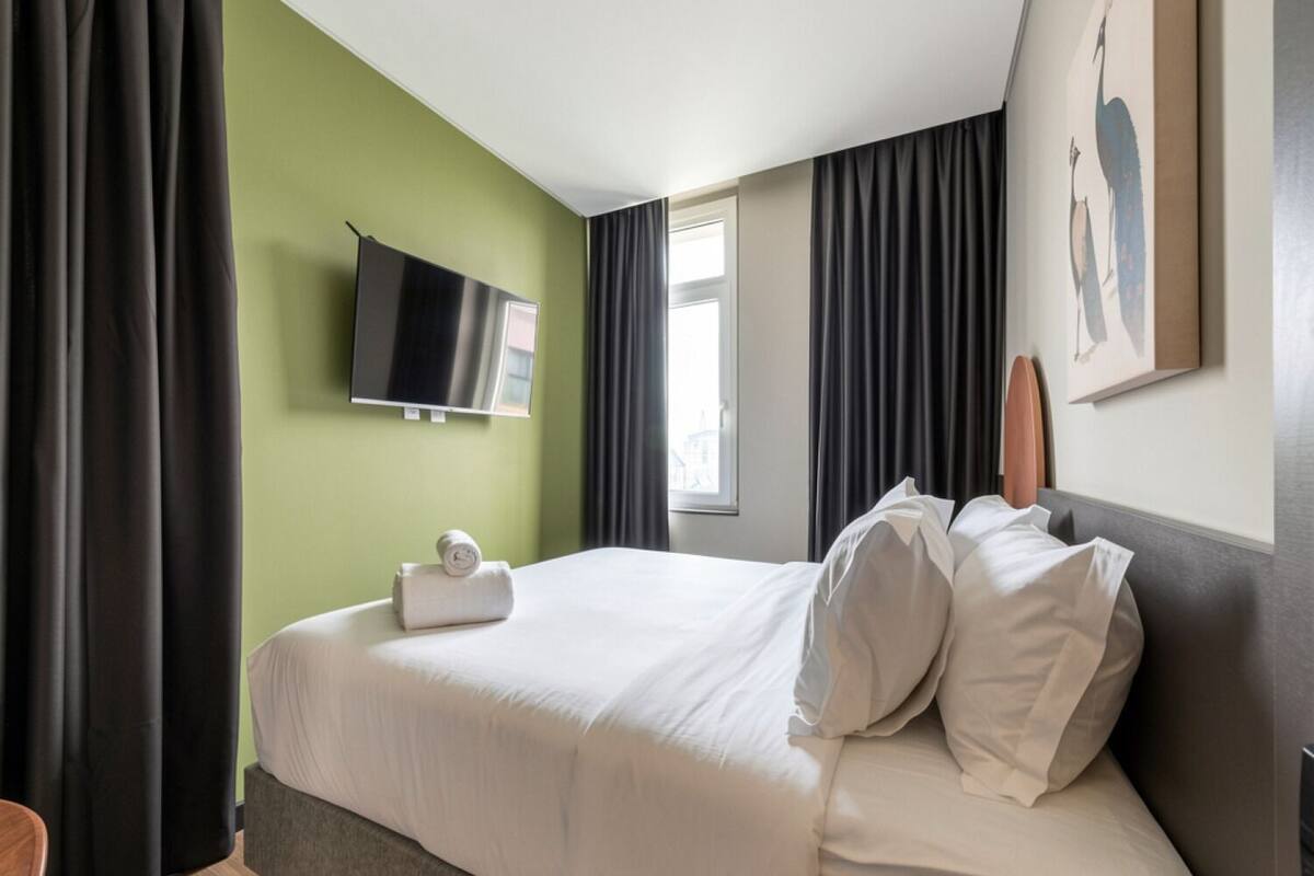 Quarto de hotel com cama, televisão de tela plana na parede, janela com cortinas e um quadro na parede.