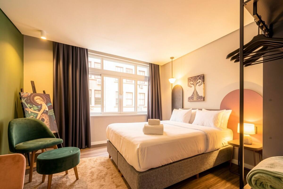 Quarto de hotel com uma cama, uma cadeira, um quadro, uma janela e um abajur.