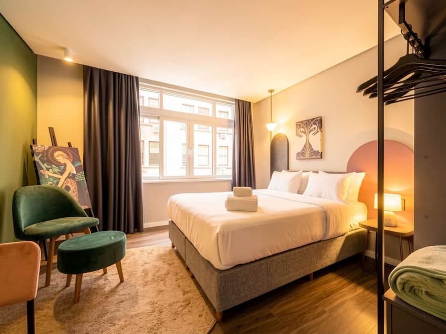 Ein Hotelzimmer mit Bett, Sessel, Gemälde, Fenster und Lampe.