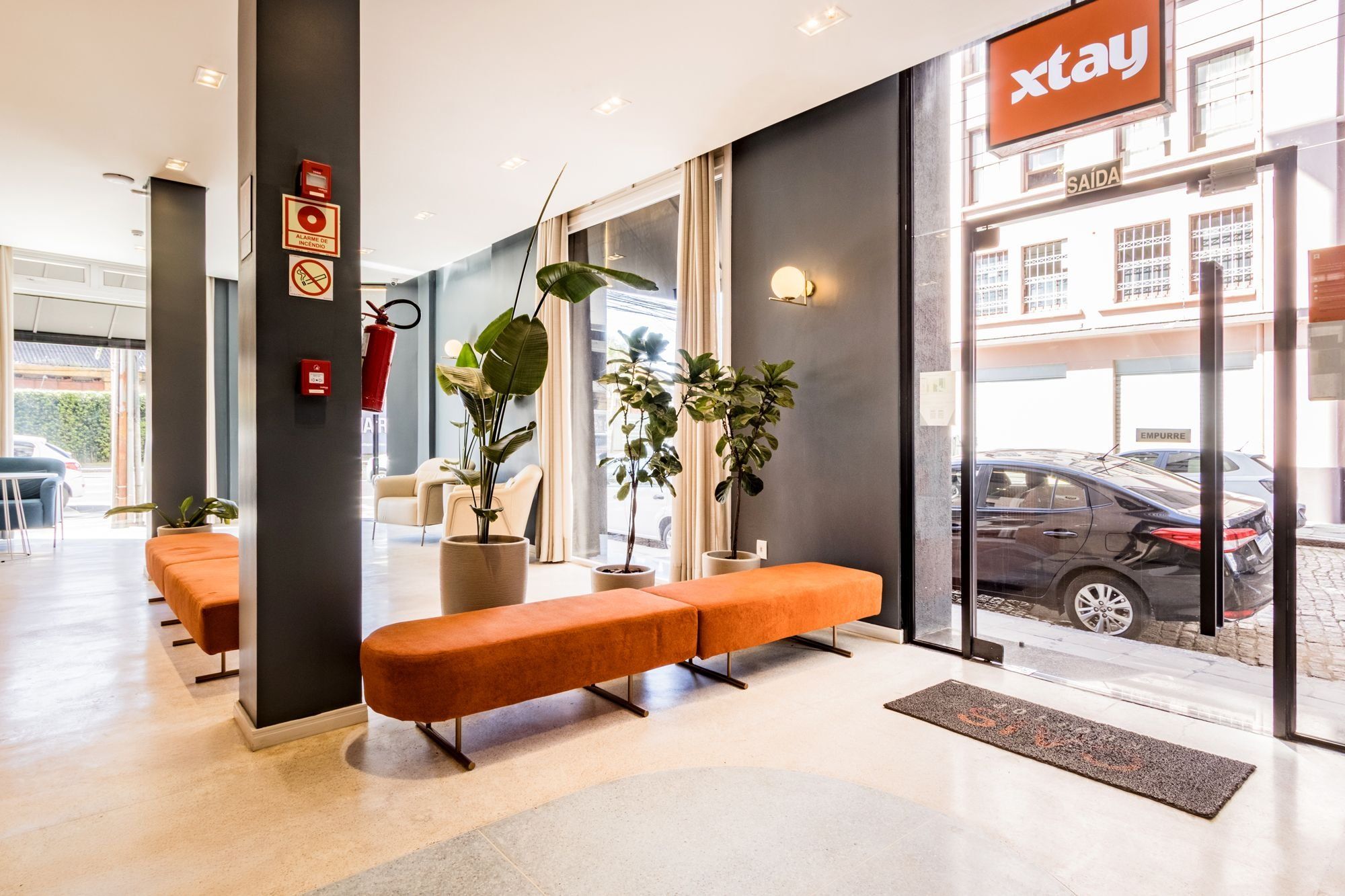 A modern hotel lobby with a reception desk, seating area, and large windows offering a view of the street.