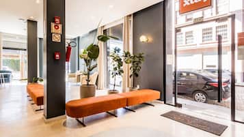 A modern hotel lobby with a reception desk, seating area, and large windows offering a view of the street.