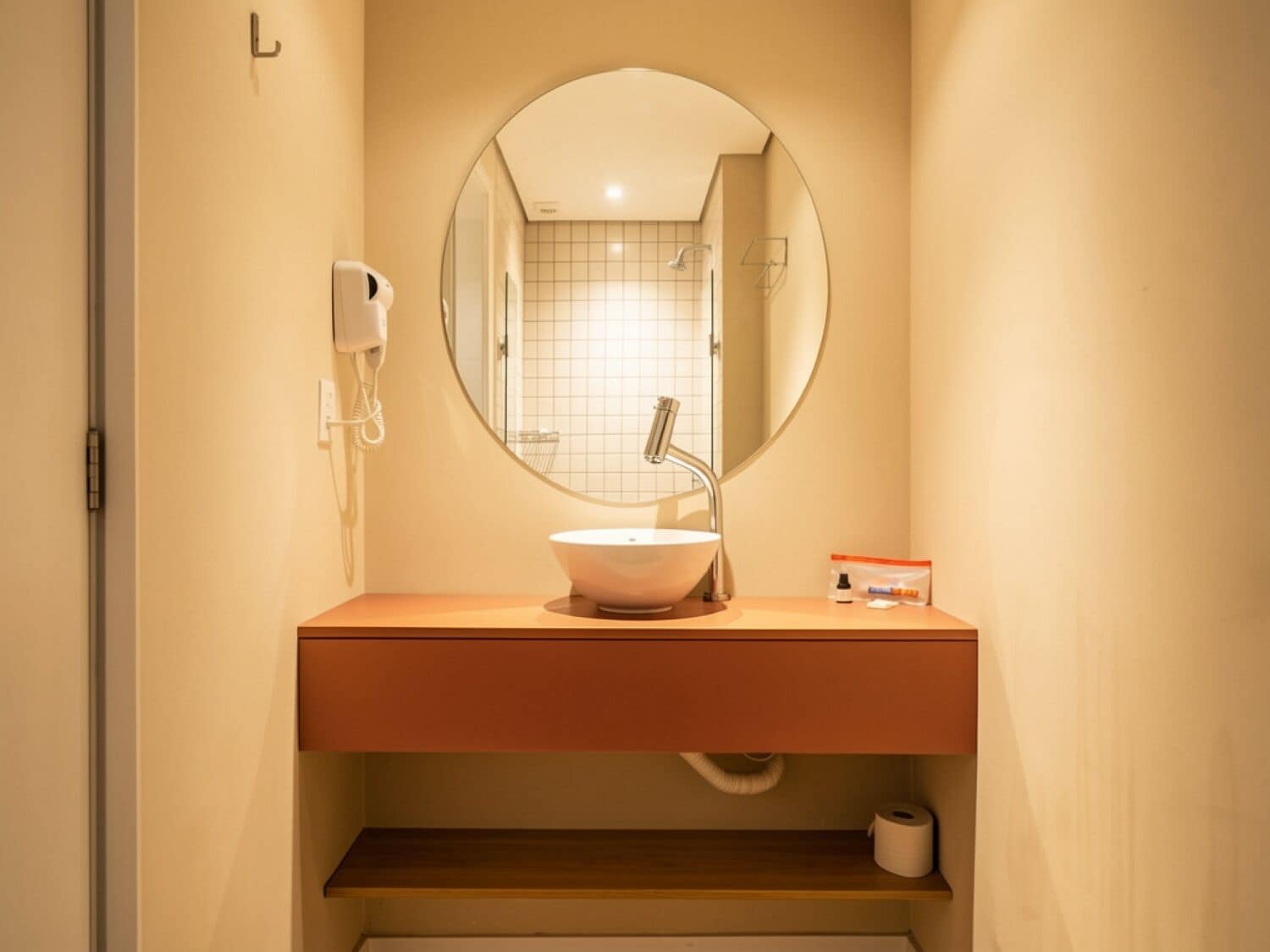 A bathroom with a round mirror, a white sink, a towel dispenser, and a toilet paper holder.