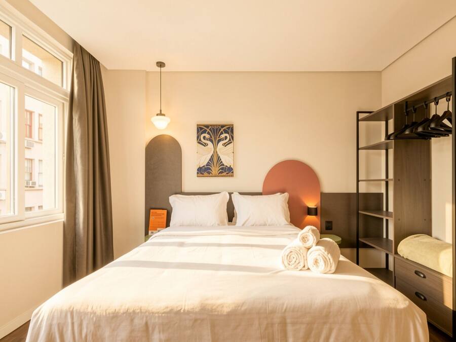 Una habitación de hotel con cama, armario, un cuadro y ventana con cortinas.