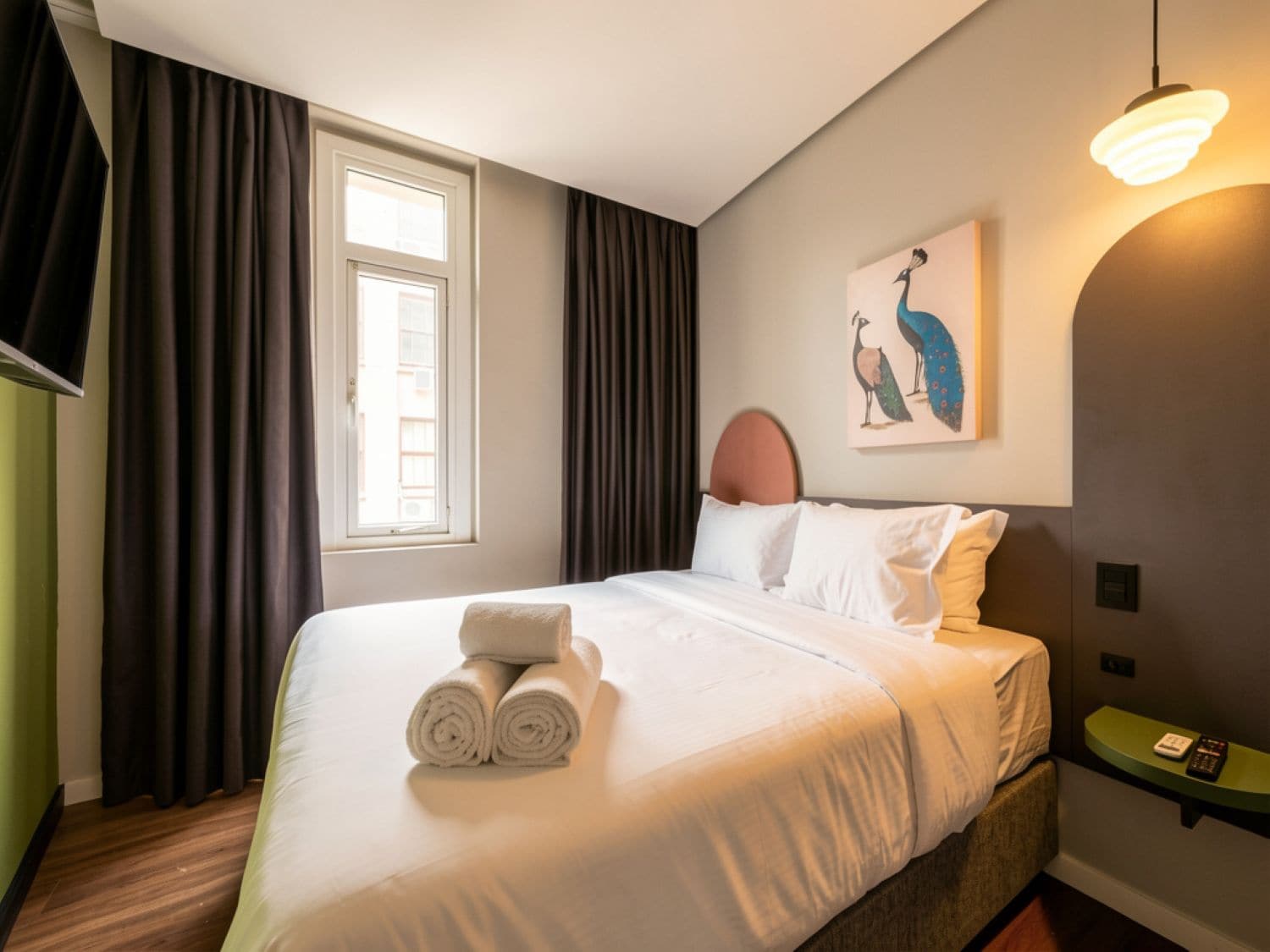 Una habitación de hotel con una cama, una ventana con cortinas, un cuadro de pájaros, una mesita de noche con un control remoto y una lámpara de pared.