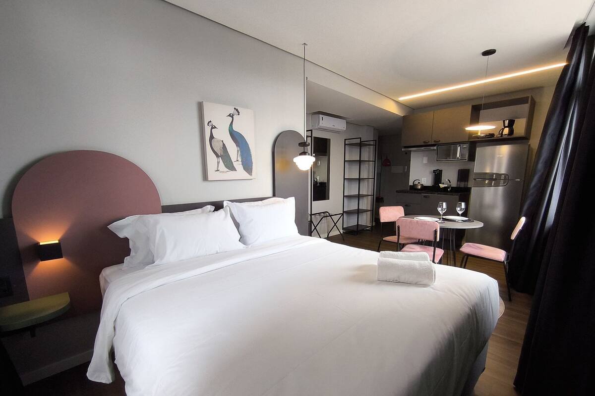 Quarto de hotel com uma cama grande, uma escrivaninha com cadeira, uma pequena cozinha e uma pintura de pavão na parede.