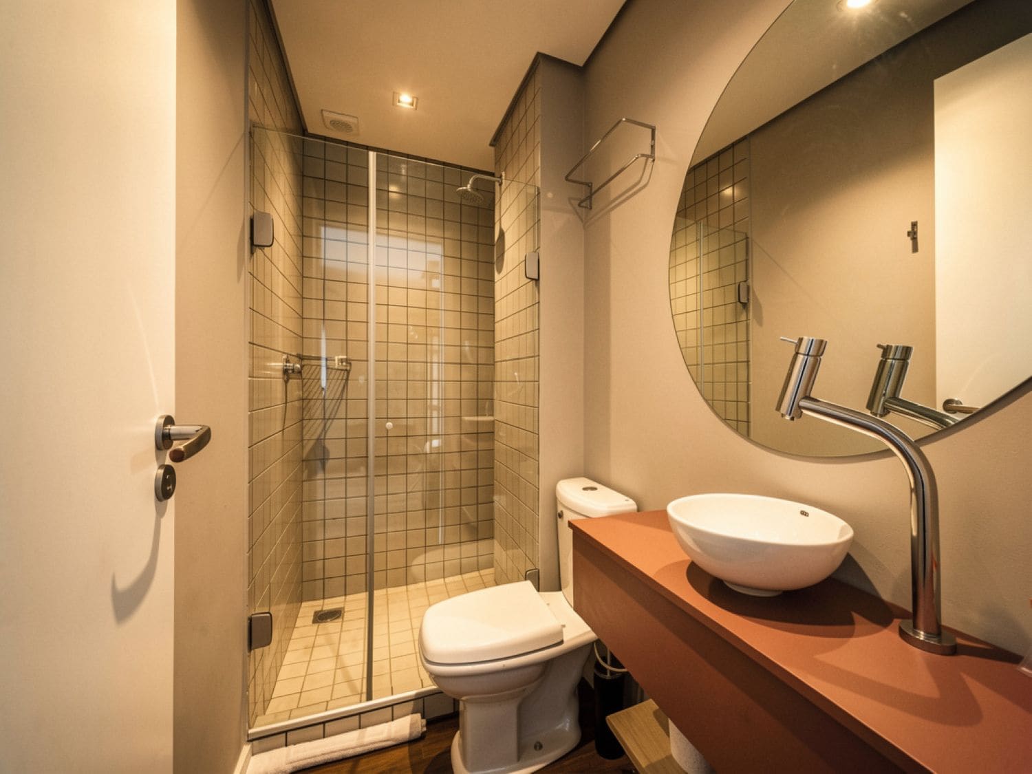 Un baño moderno con ducha de vidrio, inodoro blanco y un espejo redondo sobre un lavamanos con grifo cromado.