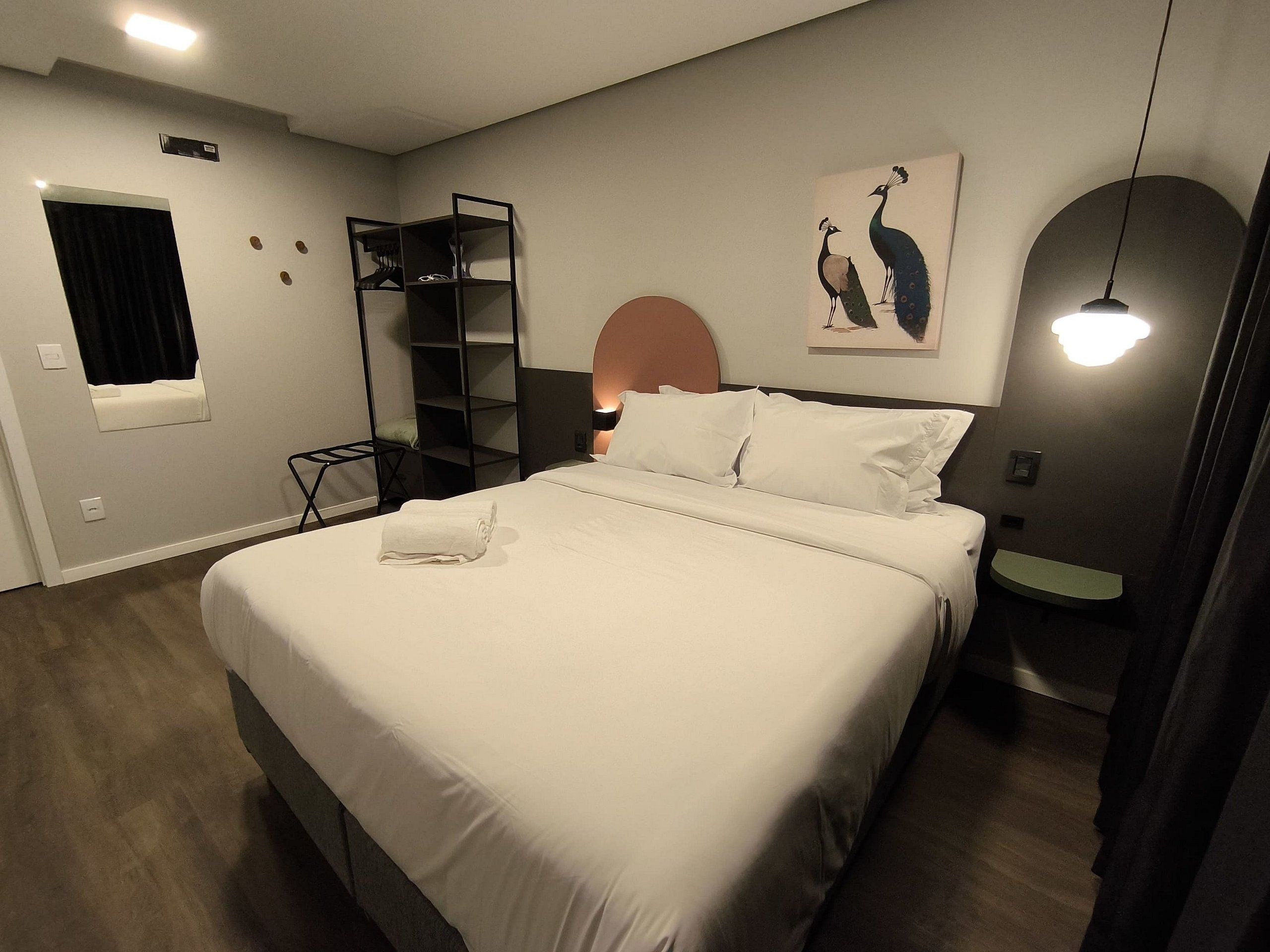 Una cama bien hecha con ropa blanca, una mesita de noche con una lámpara, un estante en la pared con ropa y un cuadro de un pavo real sobre la cama.