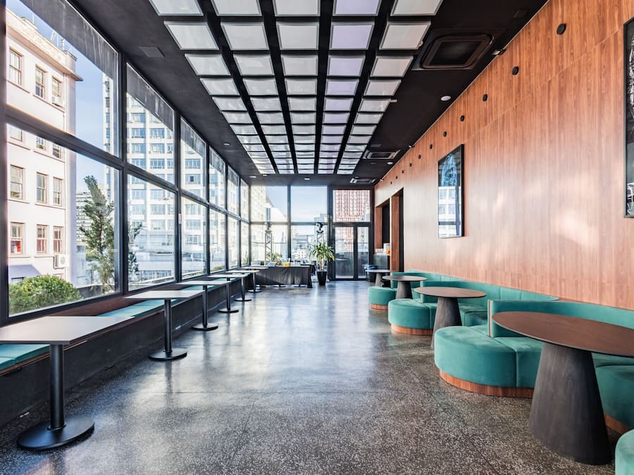 A modern interior with a long bench seating area, wooden accents, and large windows offering city views.