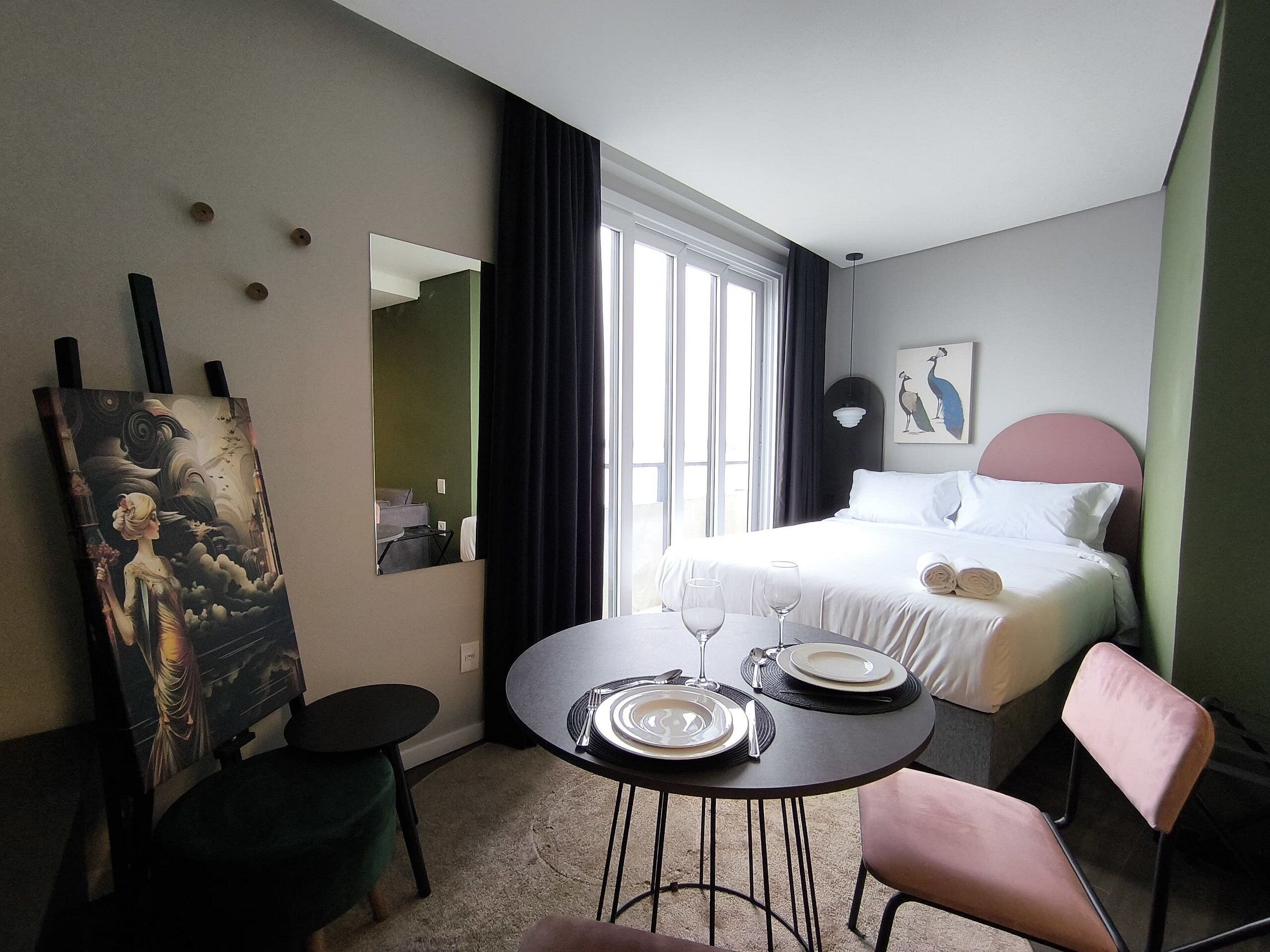 A hotel room with a bed, a dining table with plates and wine glasses, a chair, a painting on the wall, and a window with curtains.