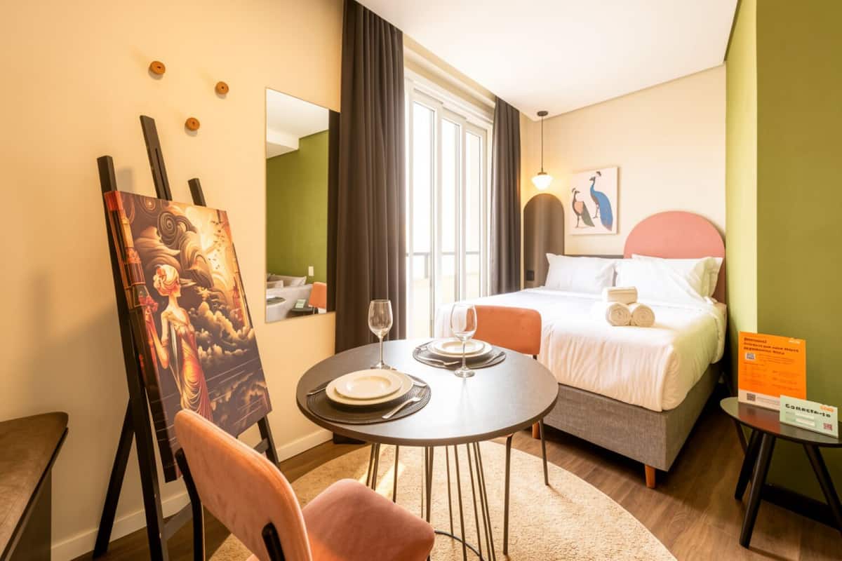 Quarto de hotel com cama, mesa de jantar com cadeiras, um quadro na parede e janela com cortinas.