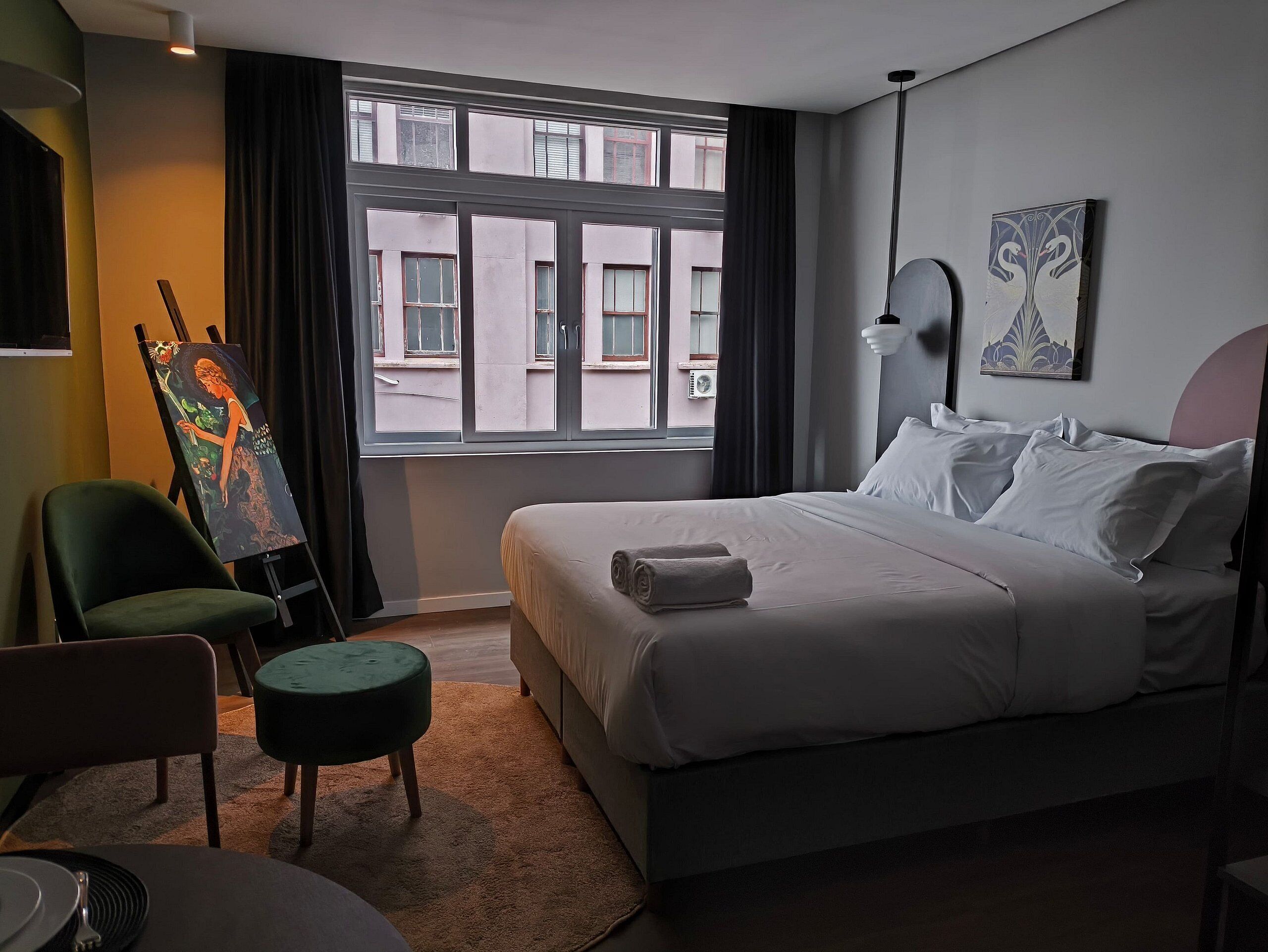 Une chambre d’hôtel avec un lit, une chaise, un tableau sur un chevalet et une fenêtre donnant sur des immeubles.