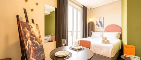 A hotel room with a bed, a dining table with chairs, a painting on the wall, and a window with curtains.