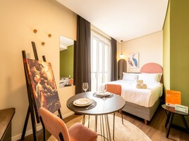 Una habitación de hotel con cama, una mesa de comedor con sillas, un cuadro en la pared y una ventana con cortinas.