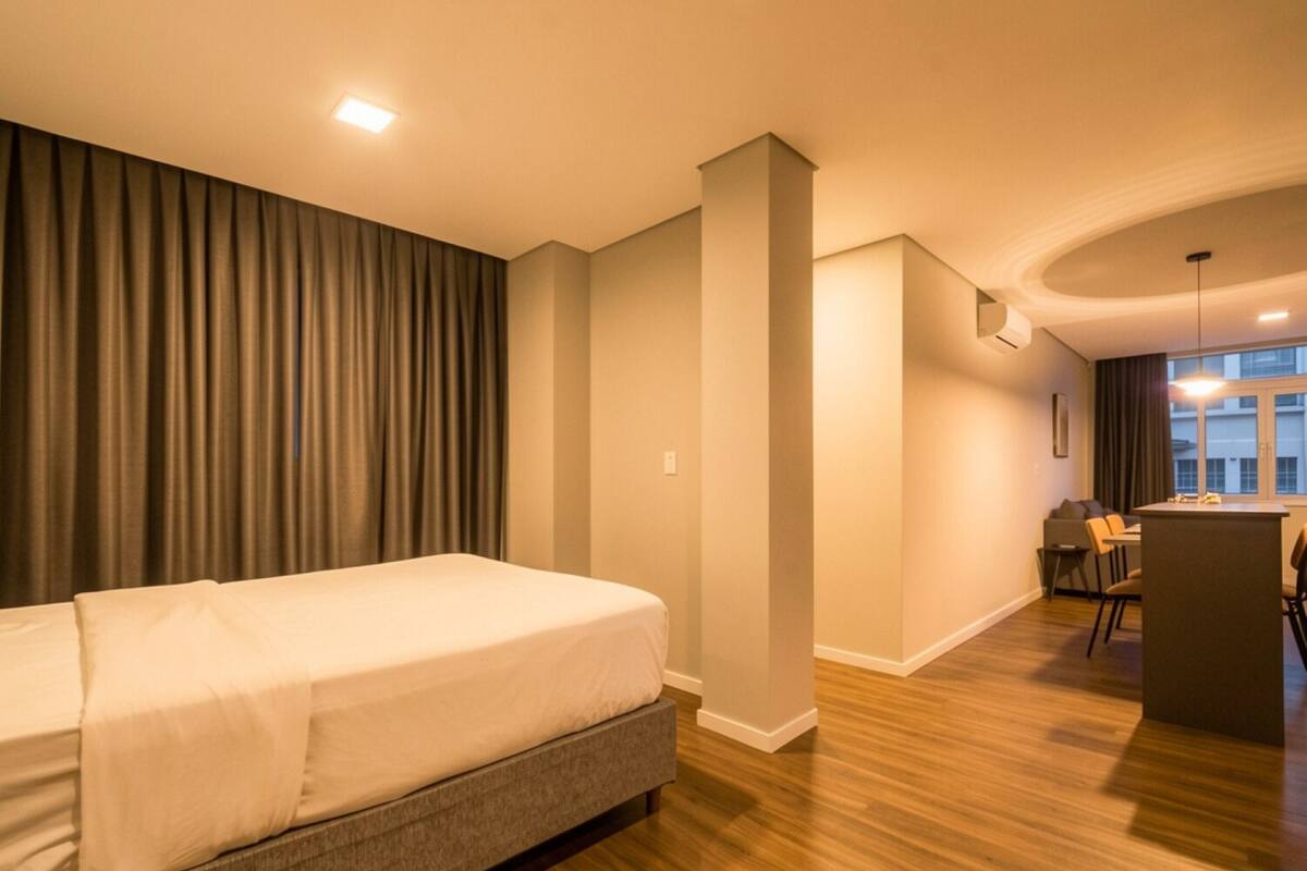 Quarto de hotel moderno com uma cama grande, uma pequena cozinha e vista para a cidade.
