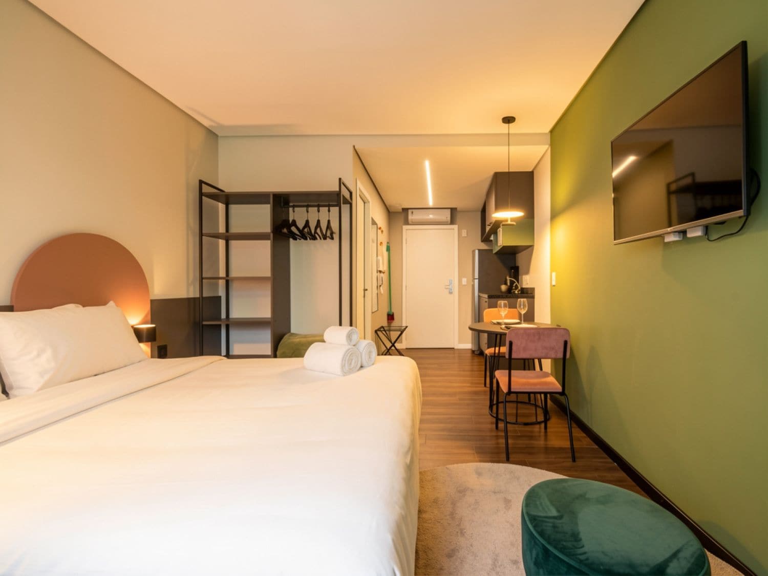 A hotel room with a bed, a desk, a chair, a mirror, and a kitchenette.