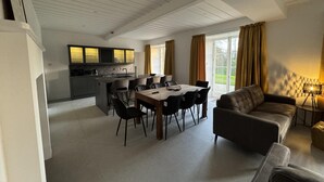 Una cucina moderna con mobili scuri e un'isola centrale, un'area pranzo con tavolo e sedie in legno, e un soggiorno con divano e lampada.