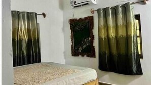 Een slaapkamer met een houten plafond, een bed met een witte matras en een geweven deken, twee gordijnen, een airconditioning en een klein decoratief spiegel.