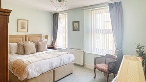 Un dormitorio con cama, una silla, una lámpara y dos ventanas con cortinas.