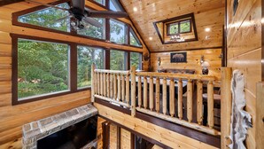 Une cabane dotée d’une cheminée en pierre, d’un balcon en bois, de grandes fenêtres et d’un ventilateur de plafond.