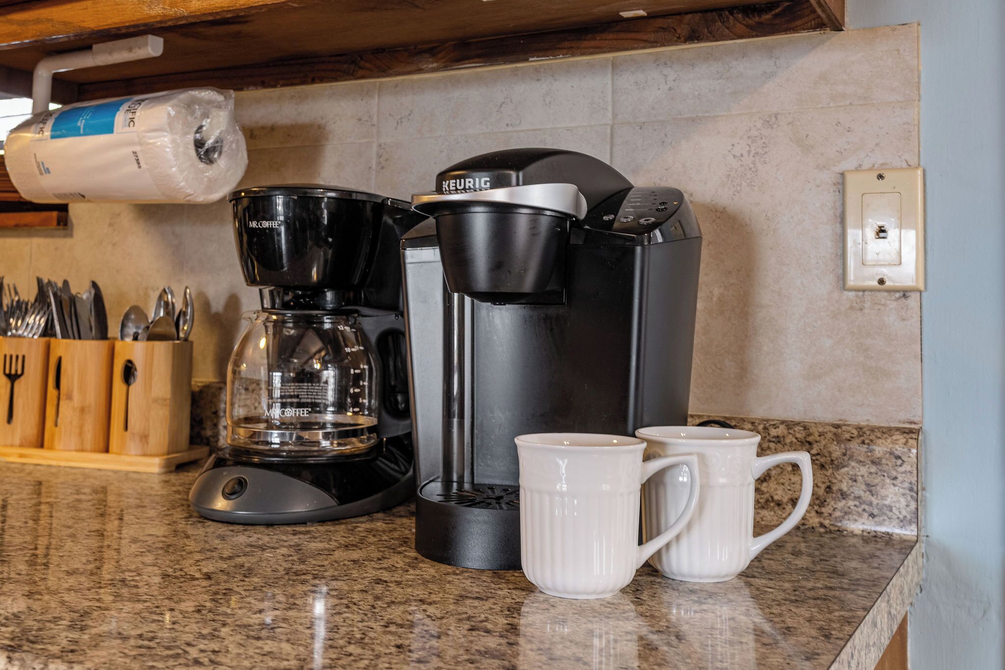 A coffee maker with two white mugs on a granite countertop.