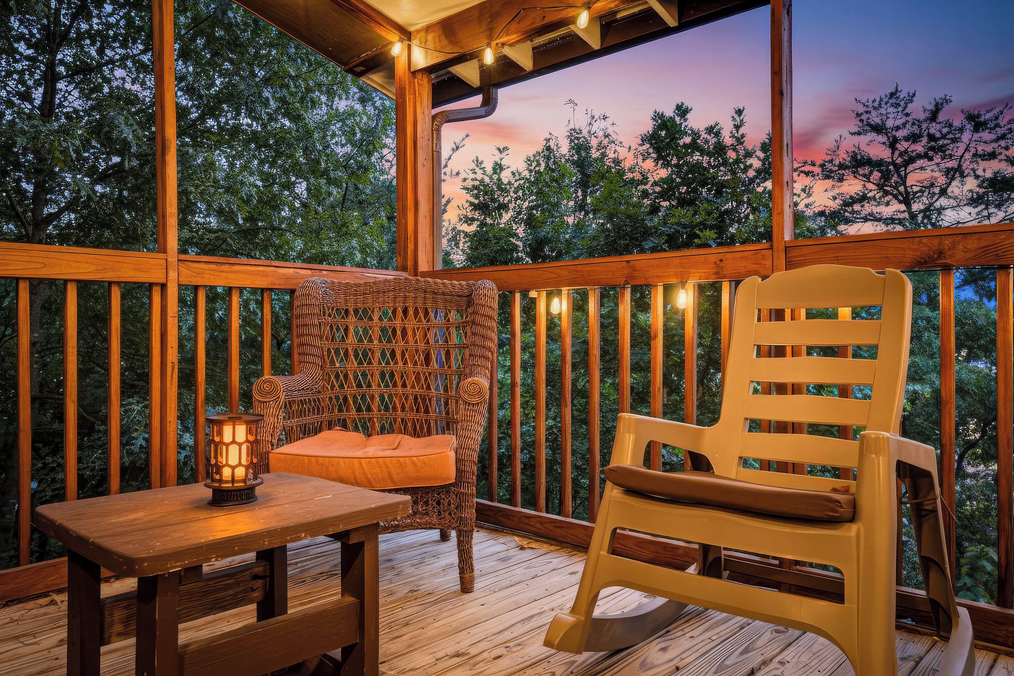 A wooden deck with a wicker chair and a wooden chair, a table with a lantern, and a view of trees at dusk.