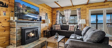Een gezellig interieur van een blokhut met een open haard, houten wanden, een bank en een televisie waarop een mooi landschap te zien is.