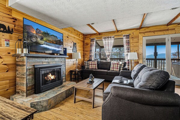 A cozy cabin interior with a fireplace, wooden walls, a sofa, and a television displaying a scenic view.