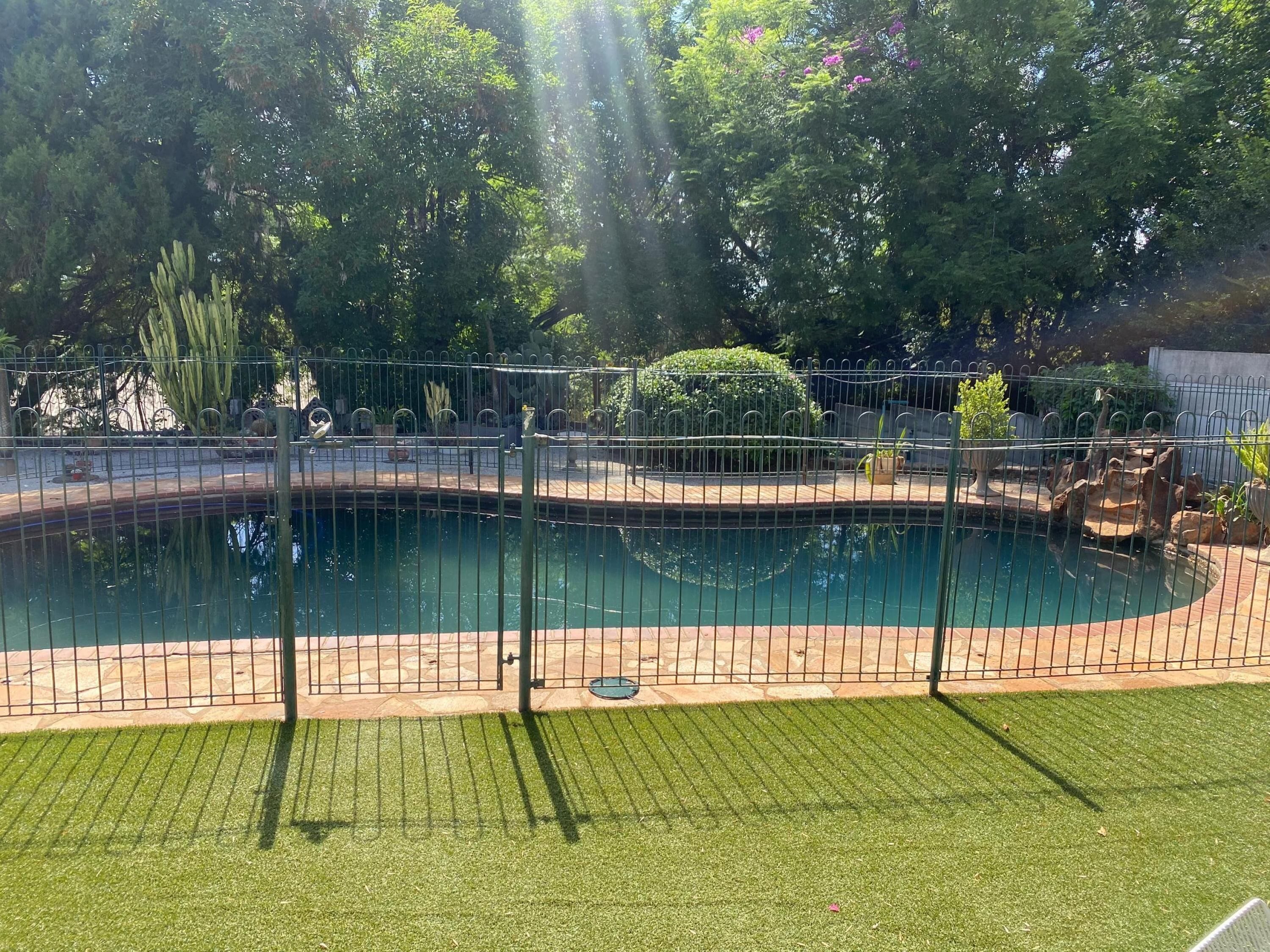 Une piscine entourée d’une clôture métallique, avec une pelouse au premier plan et des arbres en arrière-plan.