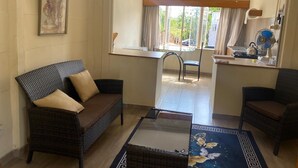 A living room with a sofa, a chair, a coffee table, and a dining area with a table and chairs. There is a window with curtains, a ceiling fan, and a painting on the wall. - Pristine 1 Bed Cottage in Northern Suburbs - 2222 (Harare)